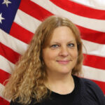 Candace Berg - Department Chairman lady with shoulder-length curly brown hair and black outfit with American flag background Photo courtesy: IceBerg Images by Candace