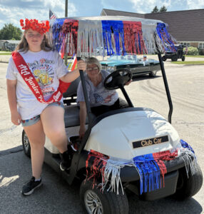 Girls wearing a poppy wreath in hair and with red sash riding a golf cart in a parade - Gackle Unit 250 Junior Poppy Queen Charlee Seigman (wearing ribbon).