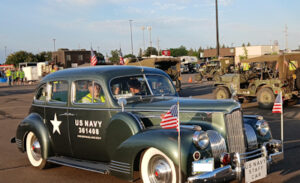 olive green 1940's era car in convoy with 2 occupants and American flags on bumper