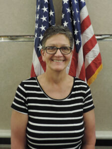 lady with short brown hair and glasses with black and white striped outfit on beige and American flag backgroundGigi Moore - Finance Board Chairman