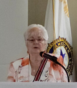 lady with silver hair and orange and white outfit talking at microphone with Auxiliary flag in background -Newly elected Department President SandySmith conducting her first leadership actions.