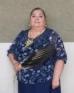 Sharon White Bear - Department Chairmanleadership@ndala.org lady with black hair, navy blue and white flowered outfit, beaded Auxiliary necklace and bird feathers on beige and cream background